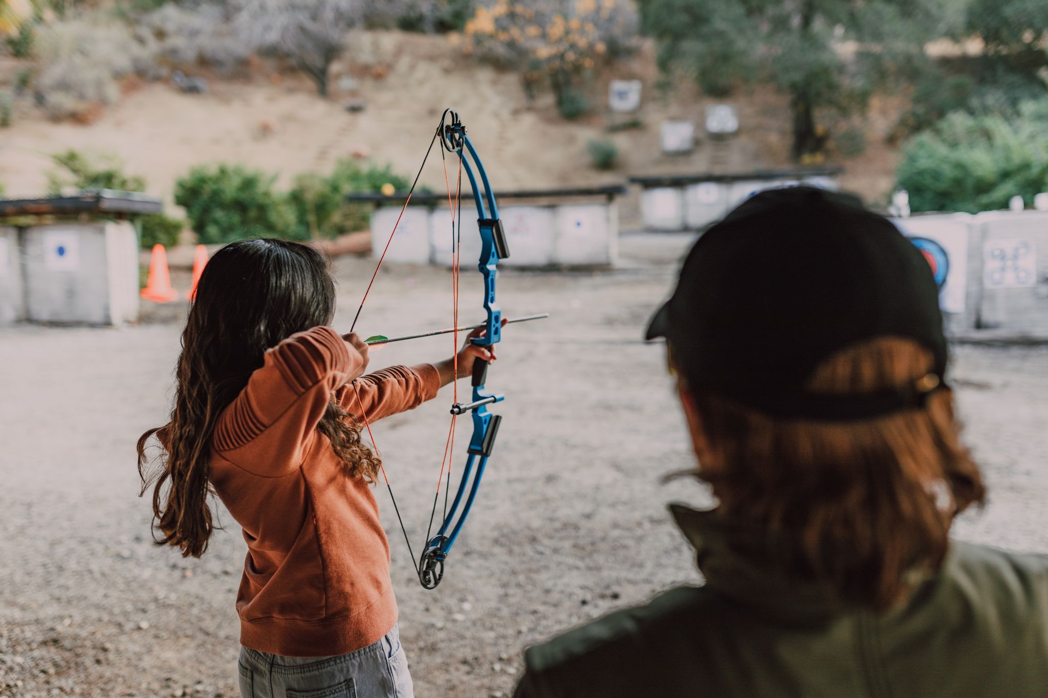 A young girl practices archery outdoors, aiming at targets with a colorful recurve bow.