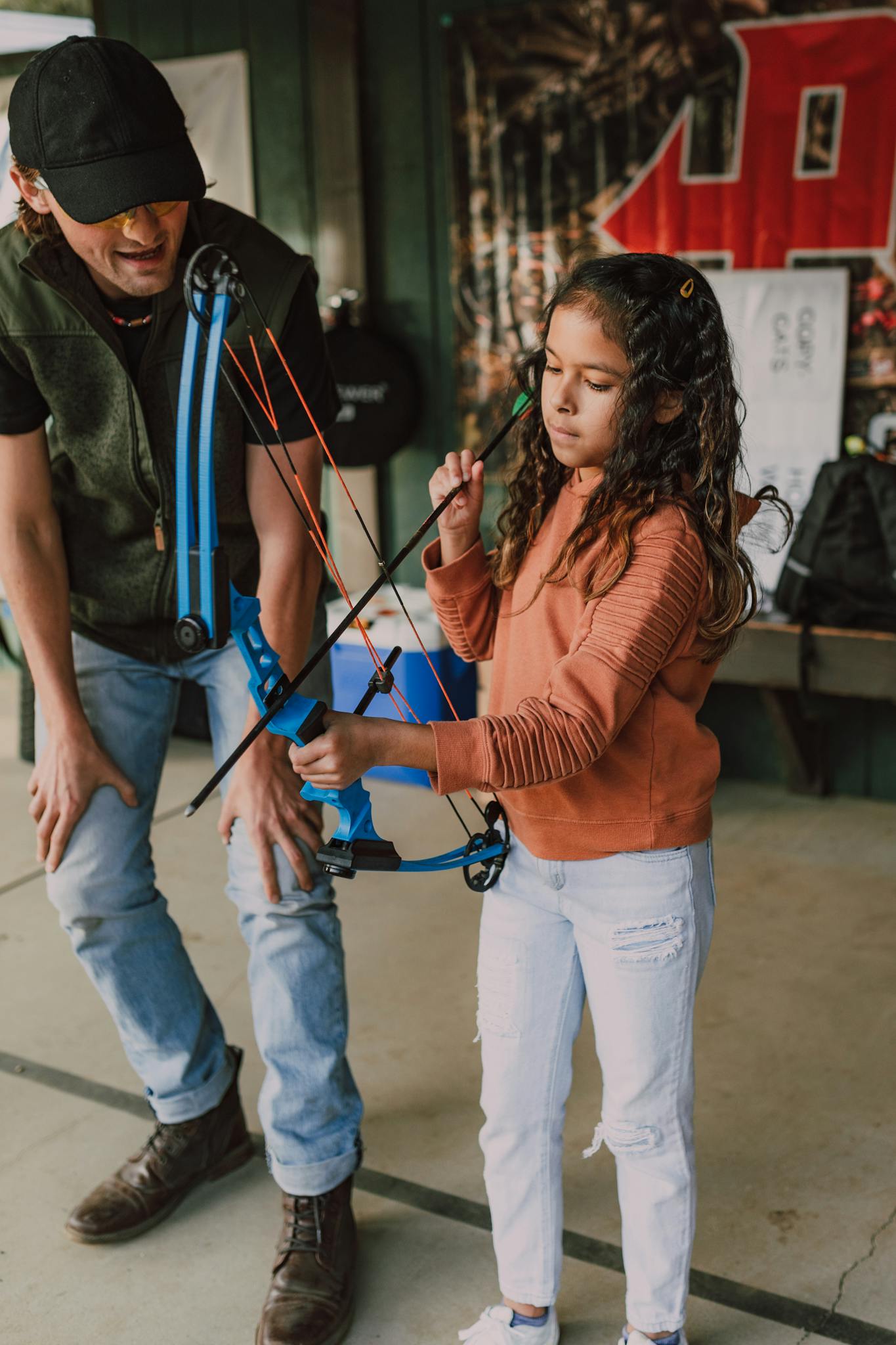 A young girl practices archery with adult guidance in an indoor setting.