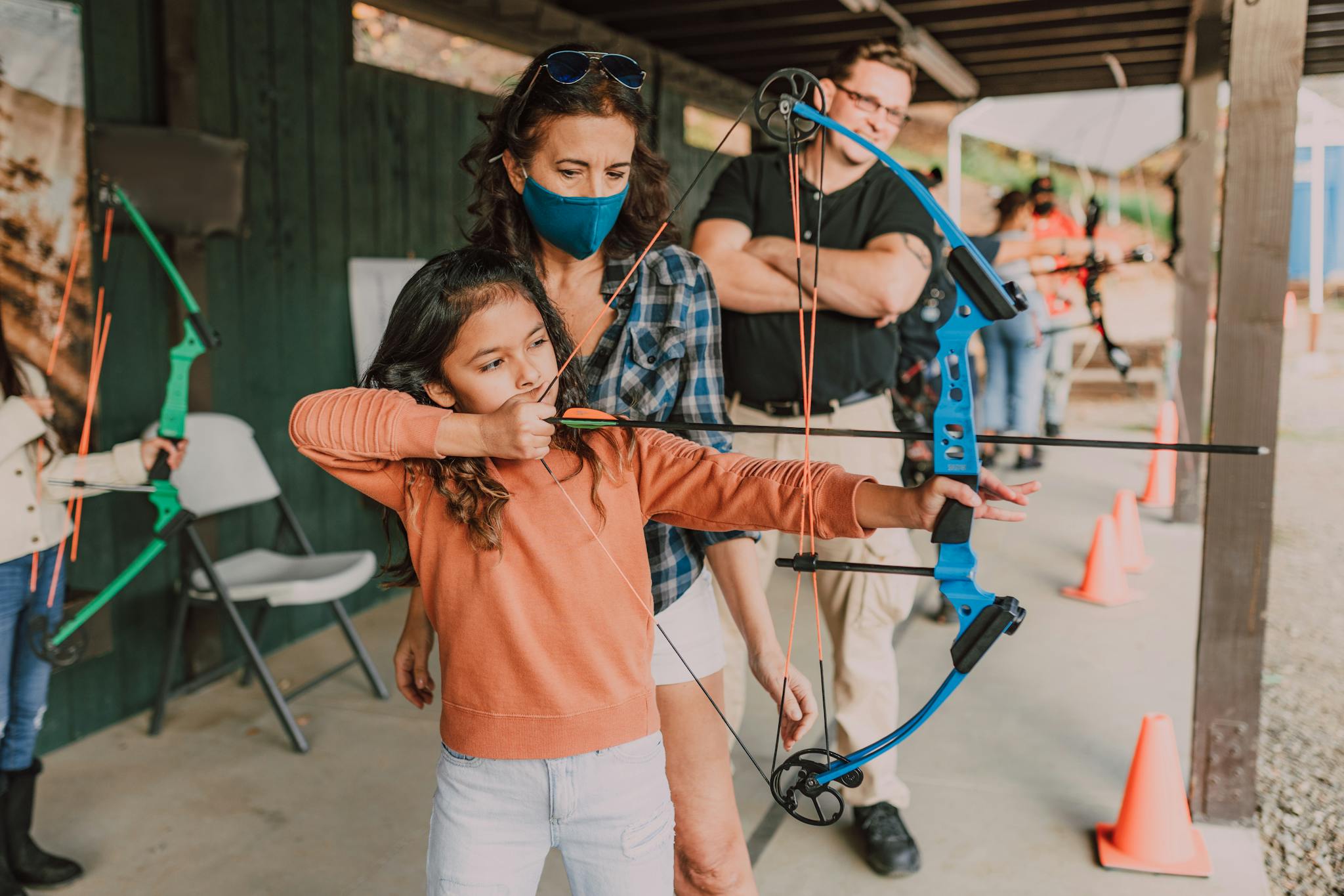A young girl receives archery lessons at an outdoor range, guided by an instructor.