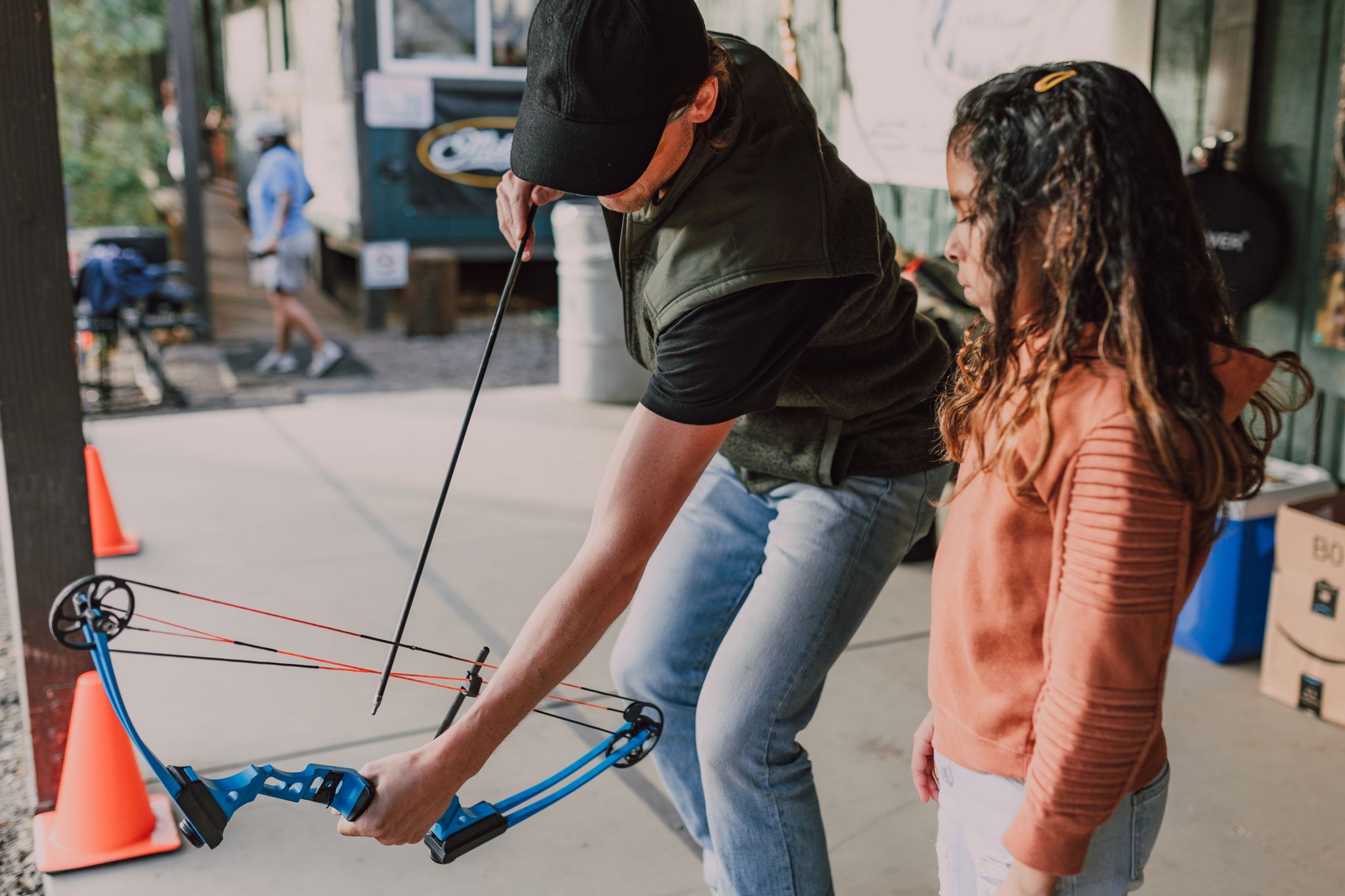 Instructor teaching a young girl archery with a compound bow at an outdoor range.