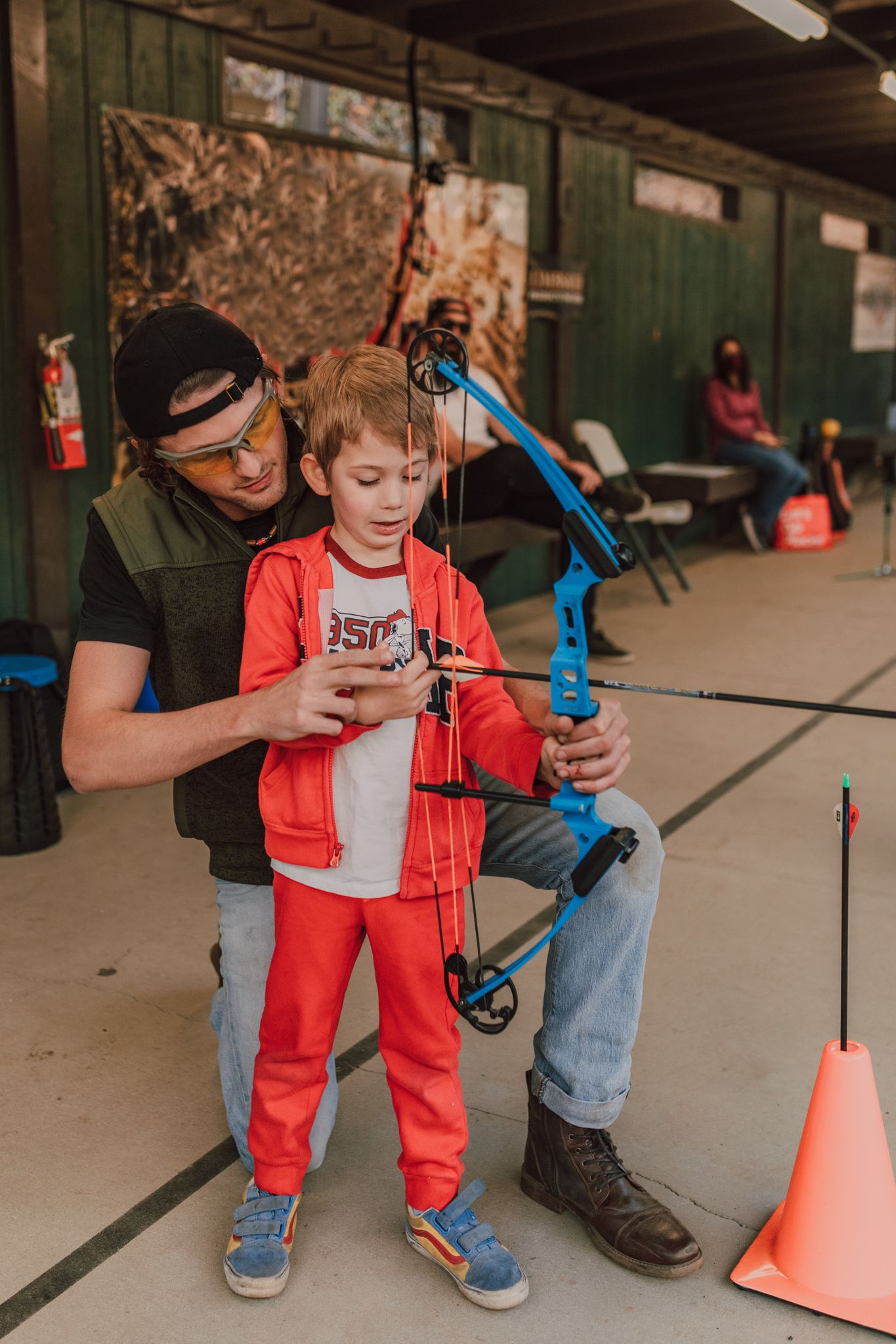 Man instructing young boy in archery at an indoor range, featuring a compound bow and safety gear.