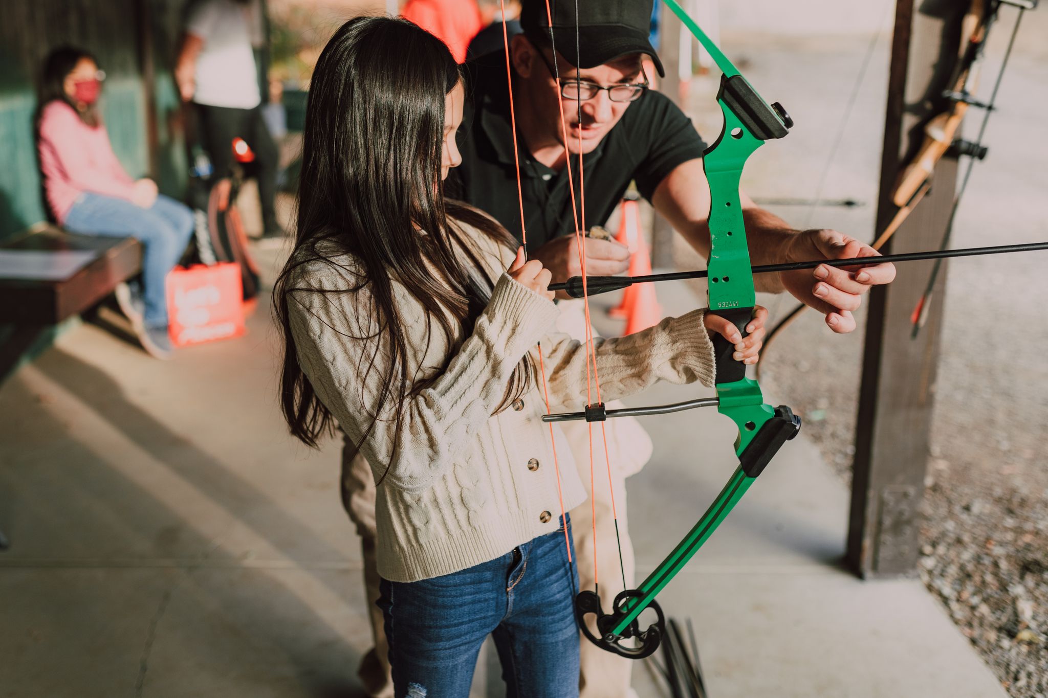 Young girl learning archery with instructor, focusing on aim outdoors.
